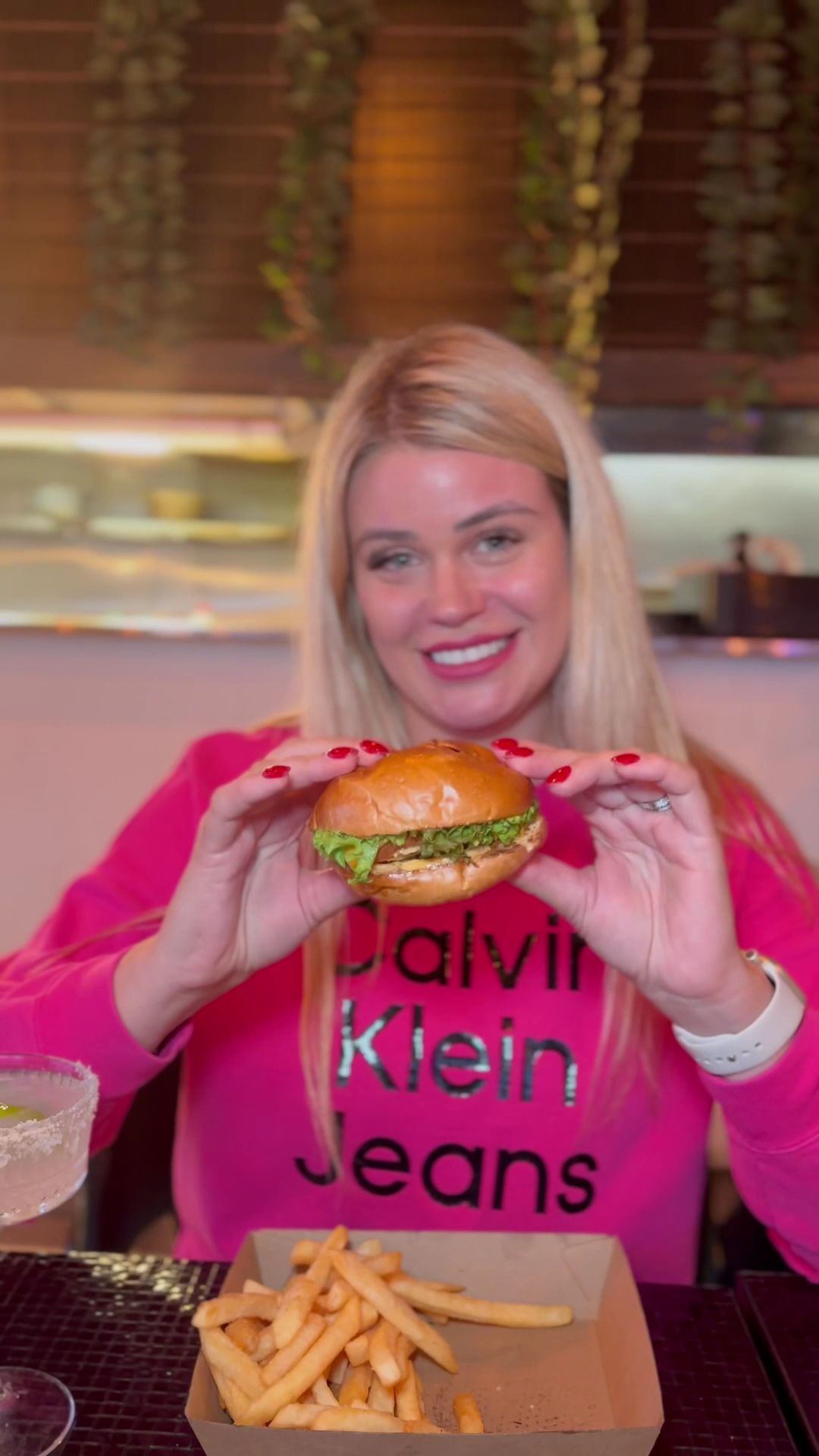 Smiling woman holding a burger with fries and a margarita cocktail at The Corner restaurant