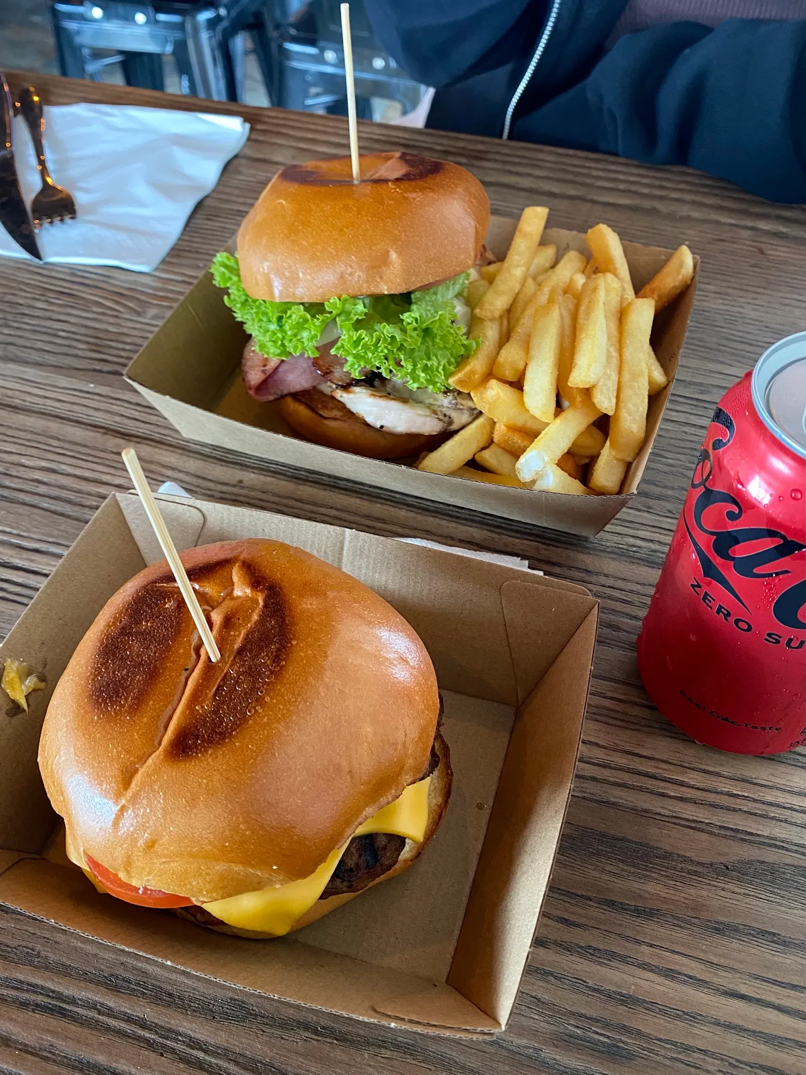 Two burgers in takeaway boxes with fries and a Coke Zero can on a dark table