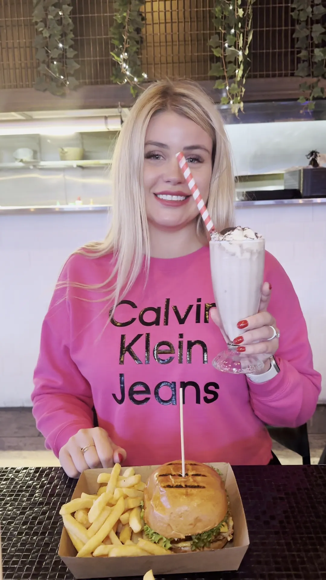 Smiling woman holding a milkshake with a burger and fries at The Corner