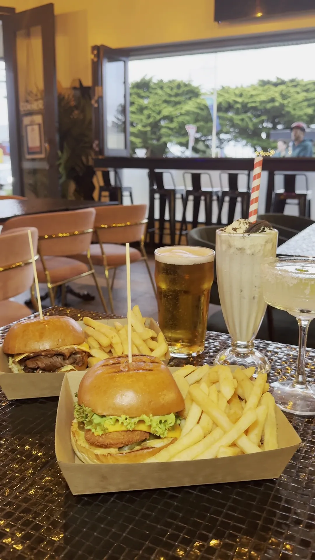 Table spread at The Corner with two burgers, chips, beer pint, milkshake and cocktail in the warm interior
